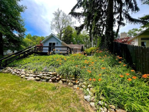 A lush garden with orange flowers and a stone border, leading up to a house surrounded by trees under a blue sky.