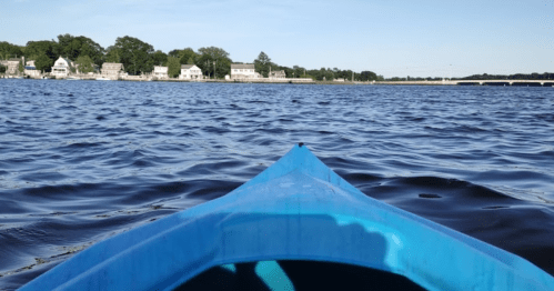 View from a blue kayak on calm water, with houses lining the shore in the background under a clear sky.
