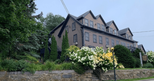A large, multi-story brown house surrounded by lush greenery and blooming white and yellow flowers.