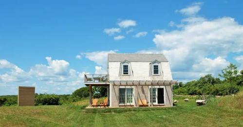 A charming two-story house with a porch, surrounded by green grass and blue skies with fluffy clouds.