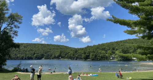 A scenic lake view with people swimming and relaxing on the shore, surrounded by green hills and fluffy clouds.