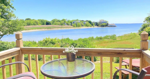 A scenic view from a wooden deck overlooking a calm lake surrounded by greenery and distant buildings.