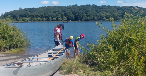 A person and two children prepare to enter a canoe by a calm lake surrounded by greenery.