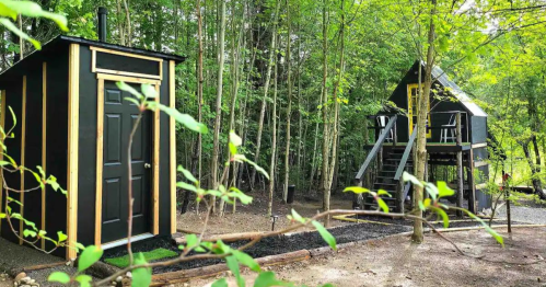Two modern cabins in a wooded area, one with a door and the other elevated on stilts with a staircase.