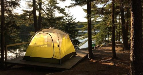 A yellow tent set up on a wooden platform by a serene lake, surrounded by tall trees and soft sunlight.