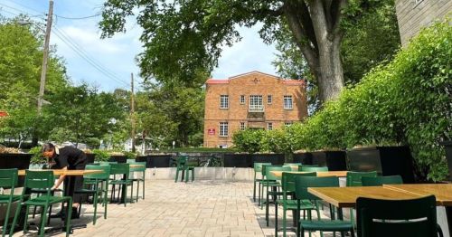 Outdoor seating area with green chairs and tables, surrounded by greenery and a brick building in the background.