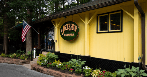A yellow ice cream shop with a sign reading "Jigger Shop," surrounded by greenery and an American flag.