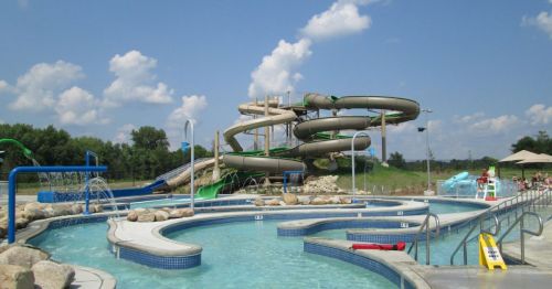 A water park featuring a winding slide, a pool with fountains, and lounge areas under a blue sky with fluffy clouds.