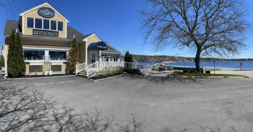 A waterfront restaurant with a yellow exterior, surrounded by trees and a clear blue sky, near a calm bay.