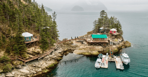 Aerial view of a coastal area with wooden structures, docks, and boats surrounded by trees and misty mountains.