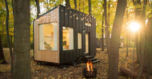 A modern tiny house with large windows, surrounded by trees and autumn foliage, with a fire pit in the foreground.
