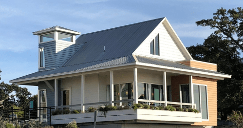 A modern two-story house with a metal roof, large windows, and a balcony, surrounded by greenery under a clear sky.
