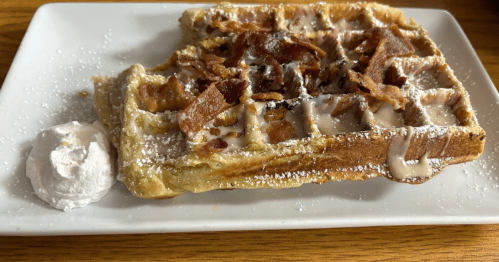 A waffle topped with crispy bacon, drizzled with syrup, and served with a dollop of whipped cream on a white plate.