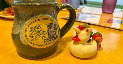 A green mug with "Sugar n' Spice Diner" logo next to a colorful rubber duck unicorn on a yellow table.