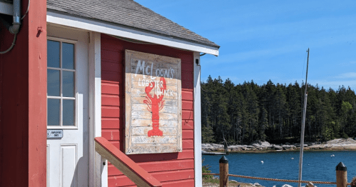 A red building with a sign reading "McLoon's Lobster Shack" near a calm waterfront and trees in the background.