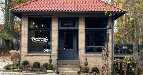 A charming coffee house with a red-tiled roof, large windows, and a welcoming "OPEN" sign. Surrounded by greenery.