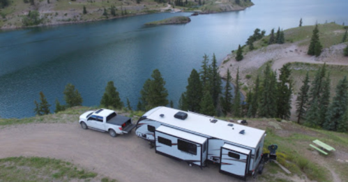 Aerial view of a camper and truck parked near a serene lake surrounded by trees and hills.