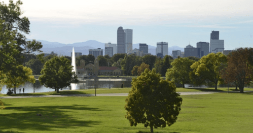 A scenic view of a city skyline with a fountain and trees in a park, set against distant mountains.