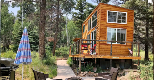 A cozy wooden cabin surrounded by trees, featuring a deck, outdoor seating, and a fire pit.