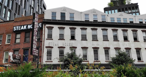Historic steakhouse building with a faded sign, surrounded by modern architecture and greenery.