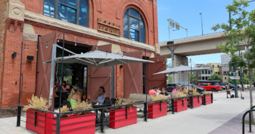 Outdoor dining area with umbrellas, people seated at tables, and a red brick building in the background.