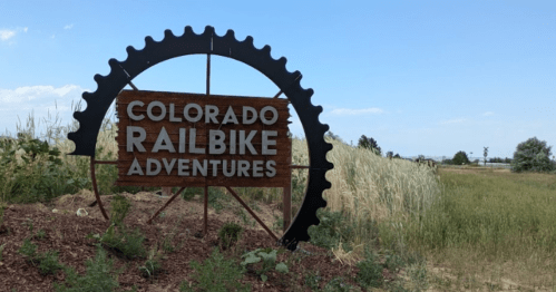 Sign for Colorado Railbike Adventures, featuring a gear design, set against a grassy landscape under a blue sky.