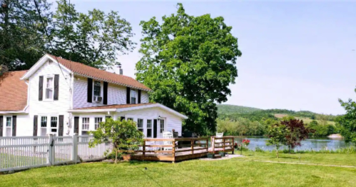 A charming white house with a deck by a serene lake, surrounded by lush greenery and rolling hills.