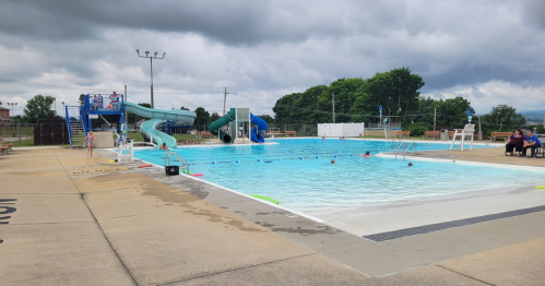 A public swimming pool with slides, cloudy skies, and people swimming and relaxing around the water.