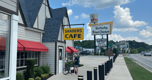 A person stands with a bicycle outside Sanders Cafe, near a Kentucky Fried Chicken sign on a sunny day.