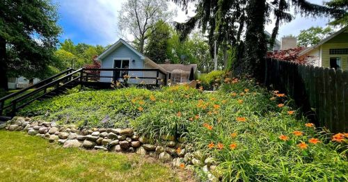 A lush garden with orange flowers and a stone wall, leading up to a house surrounded by trees and blue sky.
