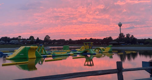 A colorful inflatable water park on a lake at sunset, with vibrant pink and orange clouds reflecting in the water.