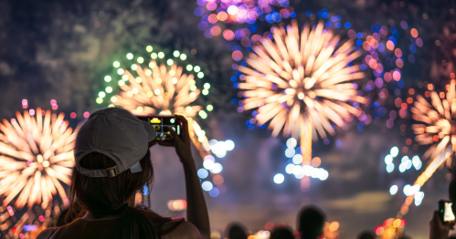 A person in a cap captures colorful fireworks on their phone against a night sky filled with vibrant bursts.