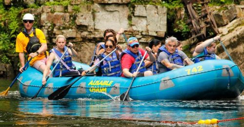 A group of people in life jackets paddling a blue raft on a river, surrounded by rocky cliffs and greenery.