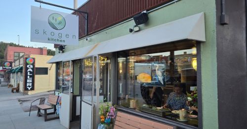 Exterior of Skogen Kitchen with a sign, outdoor seating, and a view of the street and nearby gallery.