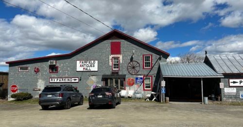 A rustic building with a sign reading "Fairfield Antiques Mall," featuring various signs and parked vehicles outside.