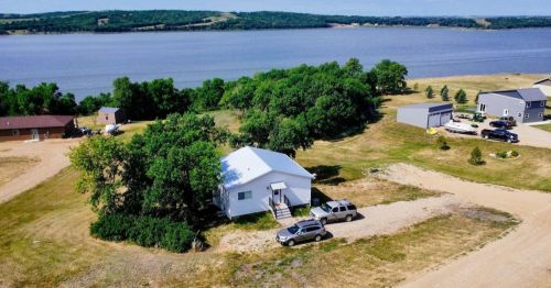 Aerial view of a white house near a lake, surrounded by trees and vehicles parked on a gravel driveway.