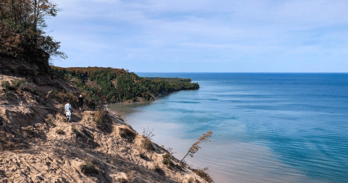 Two people walk along a sandy cliff overlooking a calm blue lake and lush green trees in the distance.