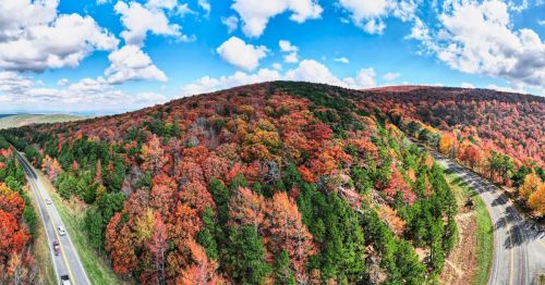 A panoramic view of a colorful autumn landscape with vibrant red and orange trees, blue skies, and a winding road.