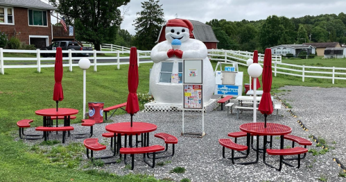 A whimsical ice cream stand shaped like a snowman, with red picnic tables and umbrellas in a grassy area.