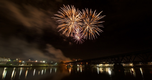 Colorful fireworks burst in the night sky above a river, with a bridge silhouetted in the foreground.