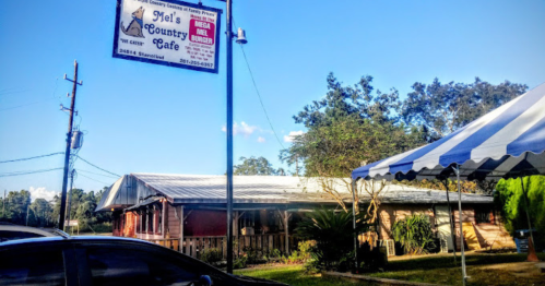 A rustic cafe building with a sign reading "Mel's Country Cafe," surrounded by trees and a blue sky.