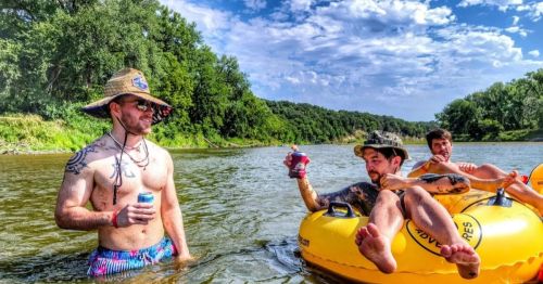 Three friends enjoy a sunny day on a river, one in a tube holding a drink, while another stands nearby with a beer.