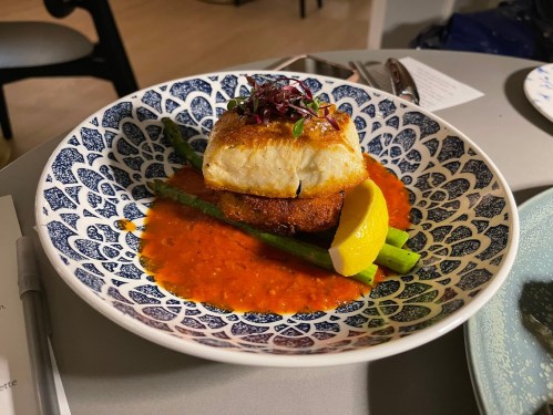 A beautifully plated dish featuring fish on a crispy base, garnished with greens and a lemon wedge, served with sauce.