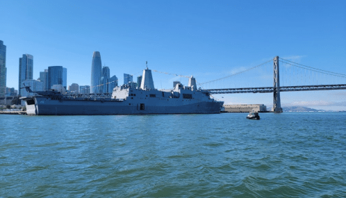A large military ship docked near a city skyline and a bridge on a clear day.