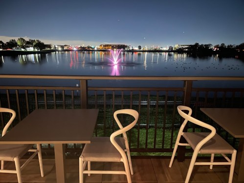 A serene lakeside view at night, featuring a colorful fountain and empty tables on a balcony.