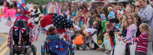 A festive parade scene with children and adults waving, dressed in patriotic attire, and decorated wheelchairs.