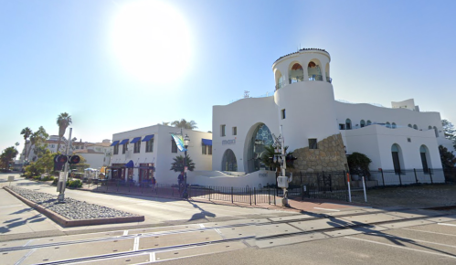A white, modern building with a tower, surrounded by palm trees and a clear blue sky, near a railway crossing.