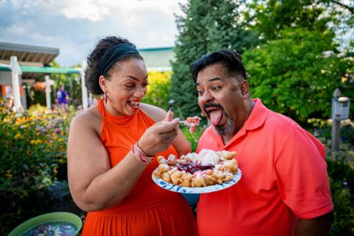 A woman in an orange dress playfully feeds a man in a pink shirt dessert from a plate in a garden setting.