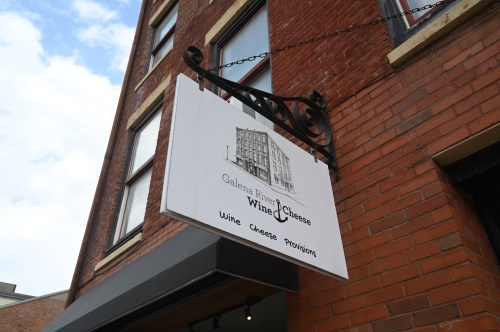 Sign for Galena River Wine & Cheese, featuring a building illustration, hanging from a brick wall.