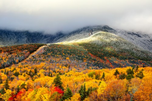 A vibrant autumn landscape with colorful trees, mountains, and clouds, showcasing a mix of fall foliage and snow.